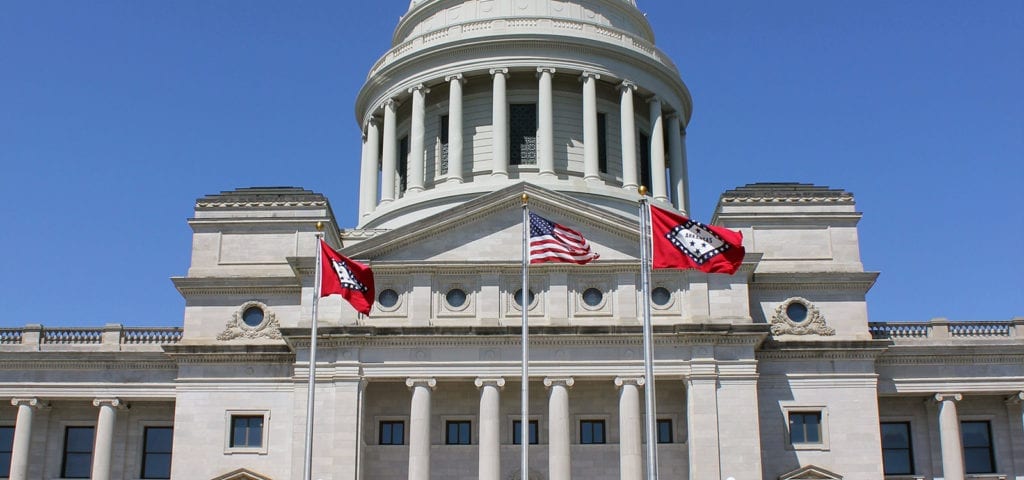 The Arkansas state flag (x2) and the U.S. flag flying in front of the Arkansas Capitol Building.