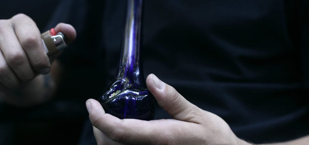 A man holds a lighter to a glass pipe loaded with ground cannabis.
