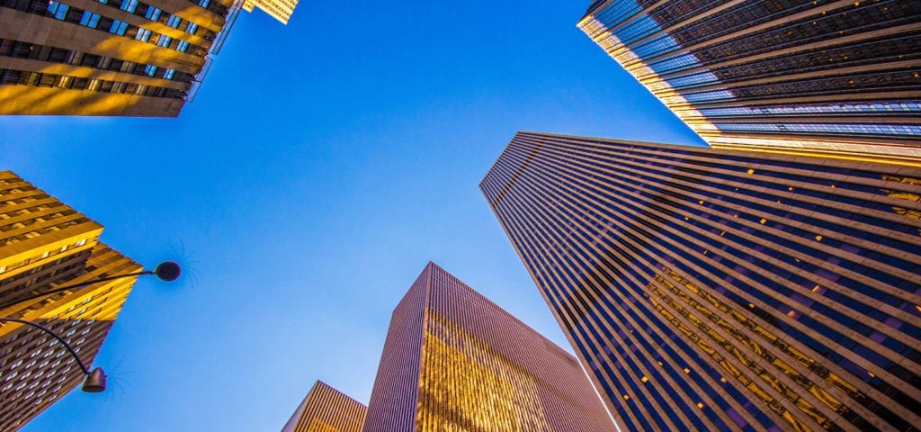 A street view in New York City, looking up between skyscrapers at clear blue sky.