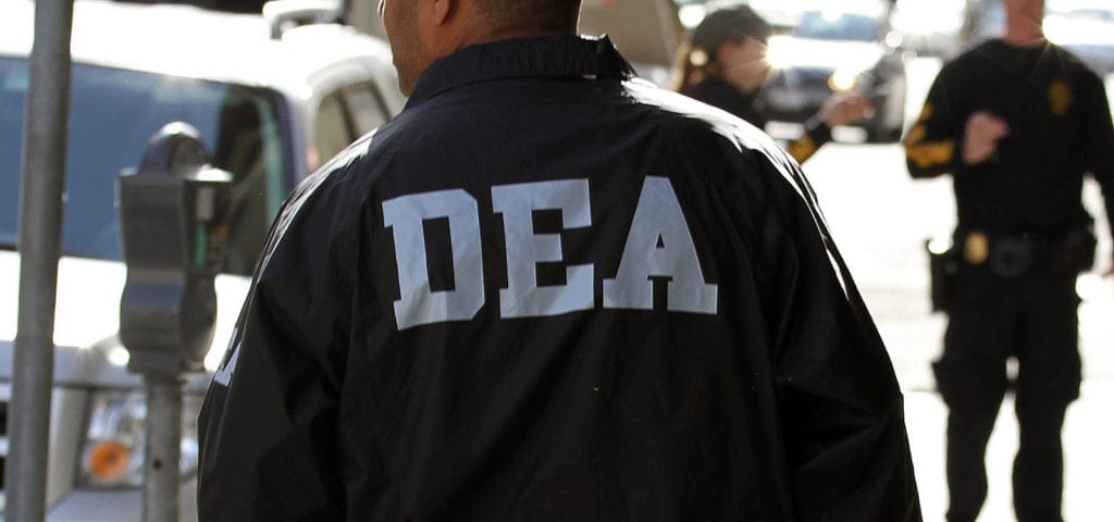 A DEA officer walking down a sidewalk next to where a raid has occurred.