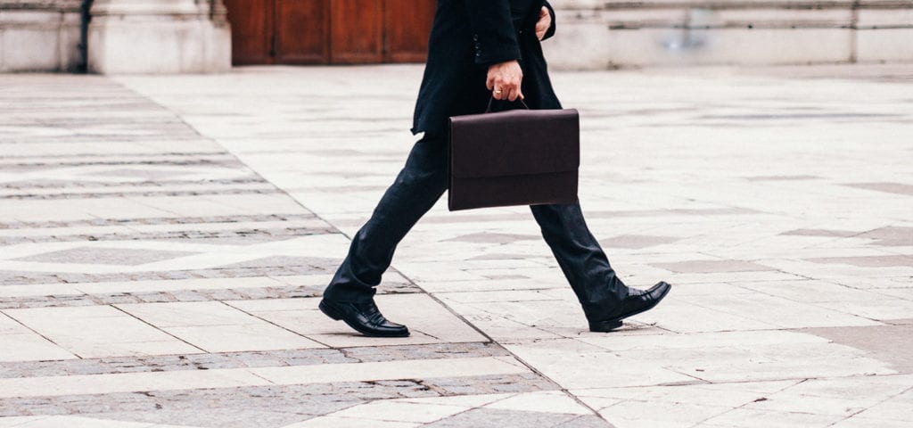 A man wearing a suit and walking across concrete while carrying a briefcase.