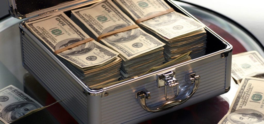 A briefcase stuffed with dollar bills sits open on a glass desk.