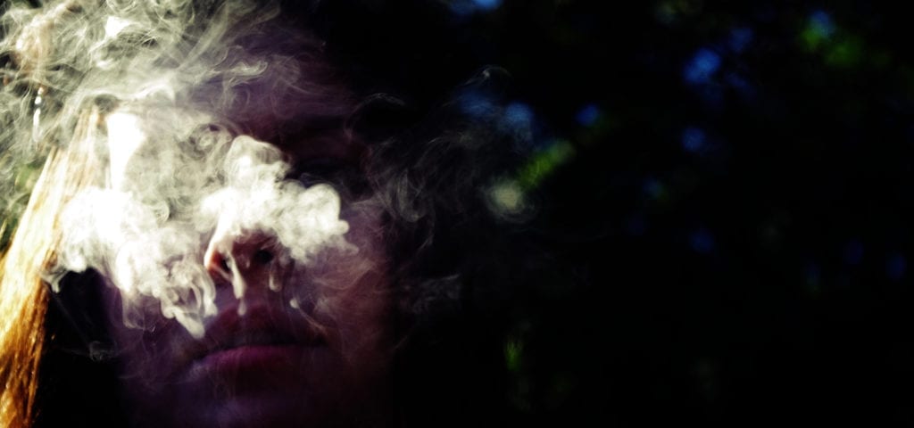 A woman exhales cannabis smoke while back-lit by the sun.