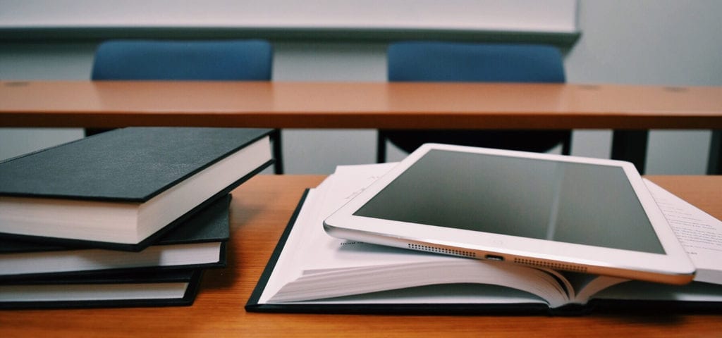 Text books and an electronic tablet sit on a desk inside of a school class room.