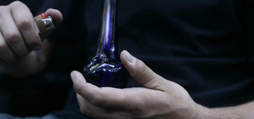 A man holds a lighter to a loaded bowl of ground cannabis.