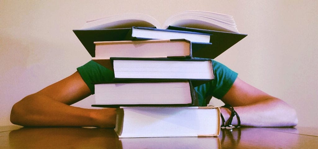 A student resting their head on a table behind a stack of large textbooks.