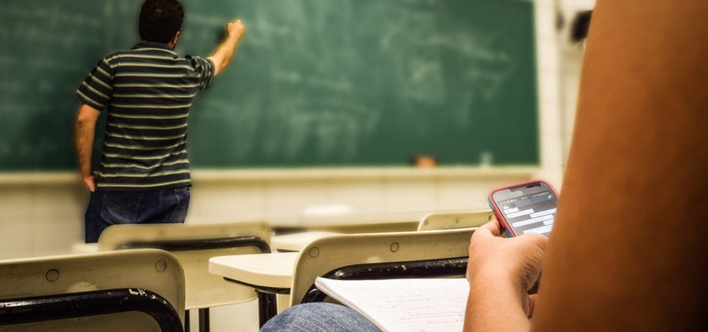 A teacher gives a lecture while one student is on their phone during class.