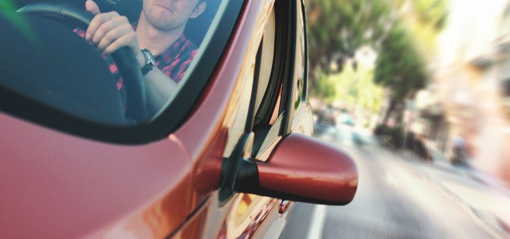 A man driving a red car down the street on a sunny day.