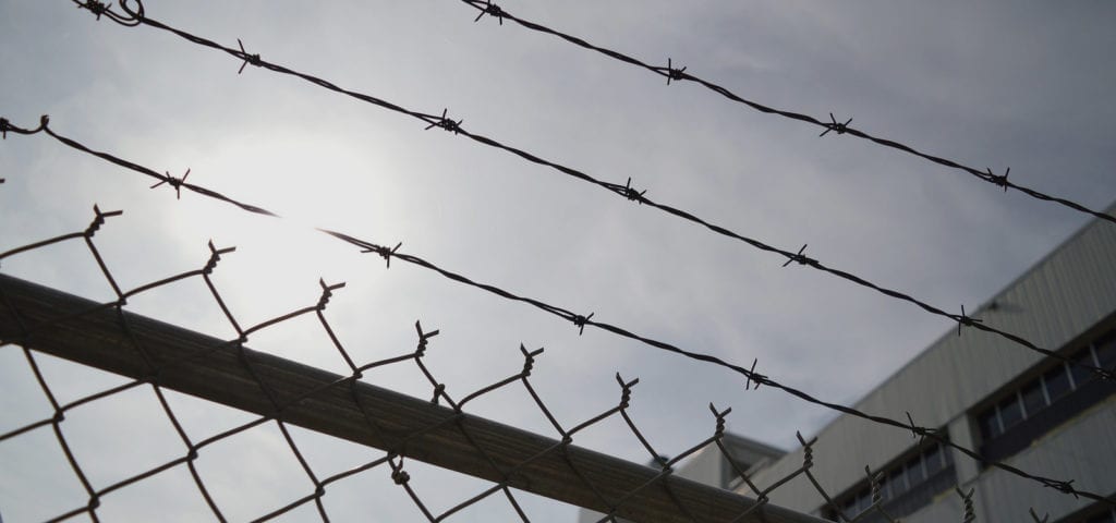 The sun peeking through clouds behind a chainlink/barbed wire fence.