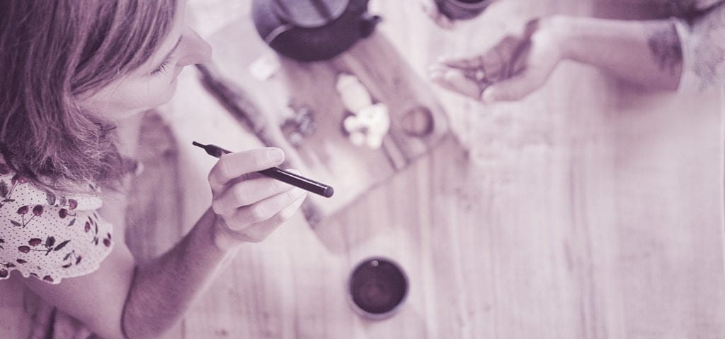 Two enthusiasts enjoy a cannabis vape pen and cups of tea at a wood-carved table.