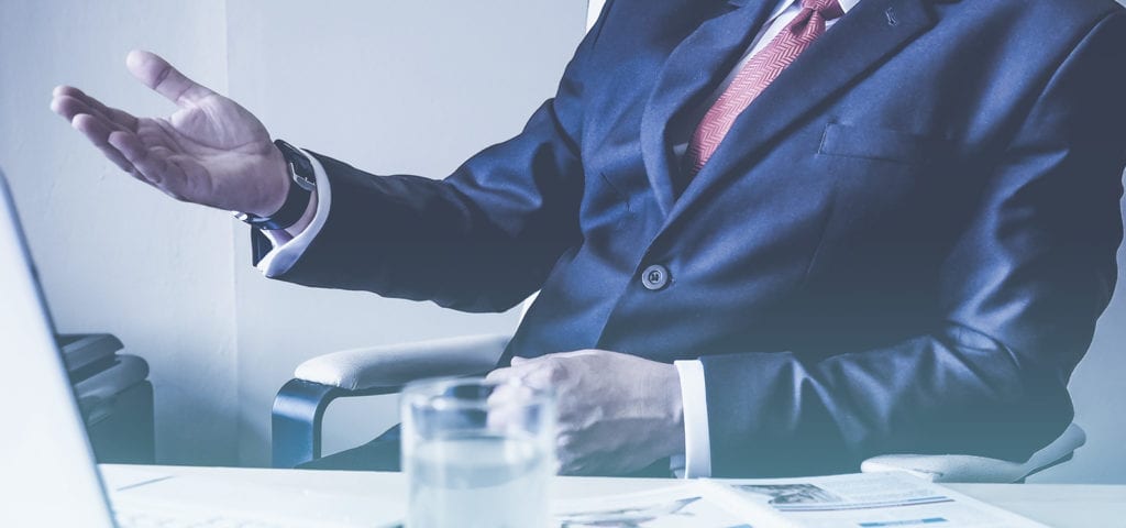 Man wearing a suit holds his hand out while sitting at a table.