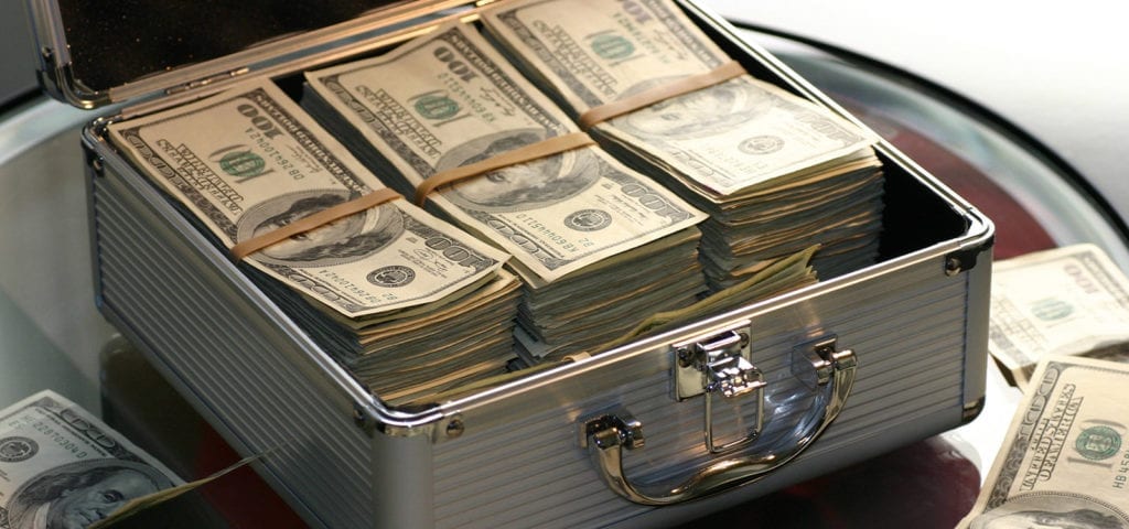 A briefcase packed full of cash sits open on a glass table, surrounded by more cash.