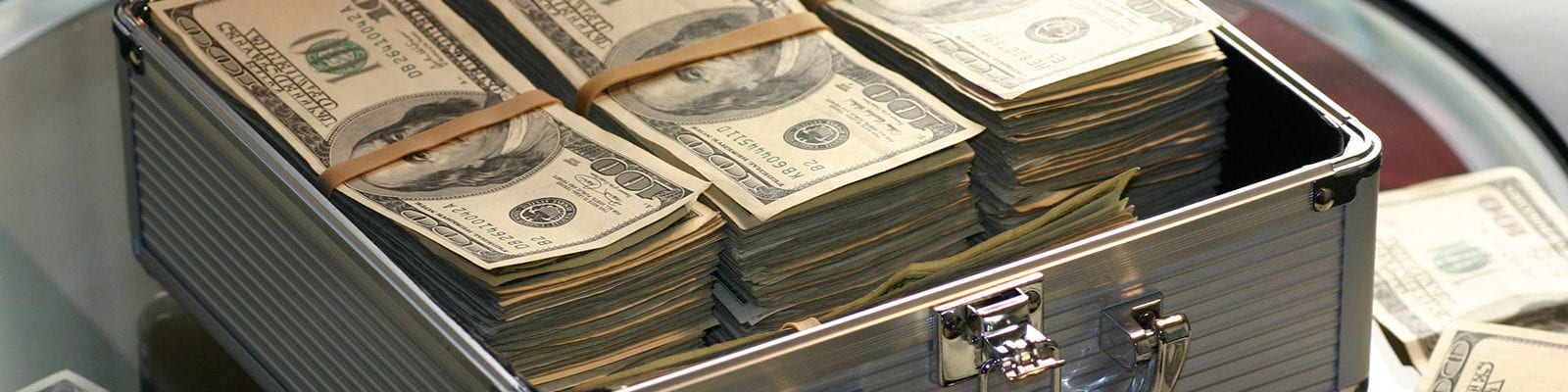 A briefcase packed full of cash sits open on a glass table, surrounded by more cash.