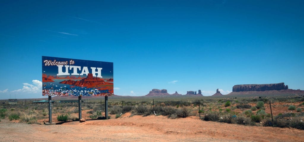 A "Welcome to Utah" sign alongside the road with several sandstone monuments on the horizon behind it.