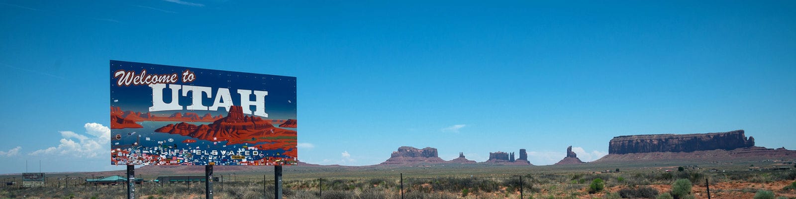 A "Welcome to Utah" sign alongside the road with several sandstone monuments on the horizon behind it.