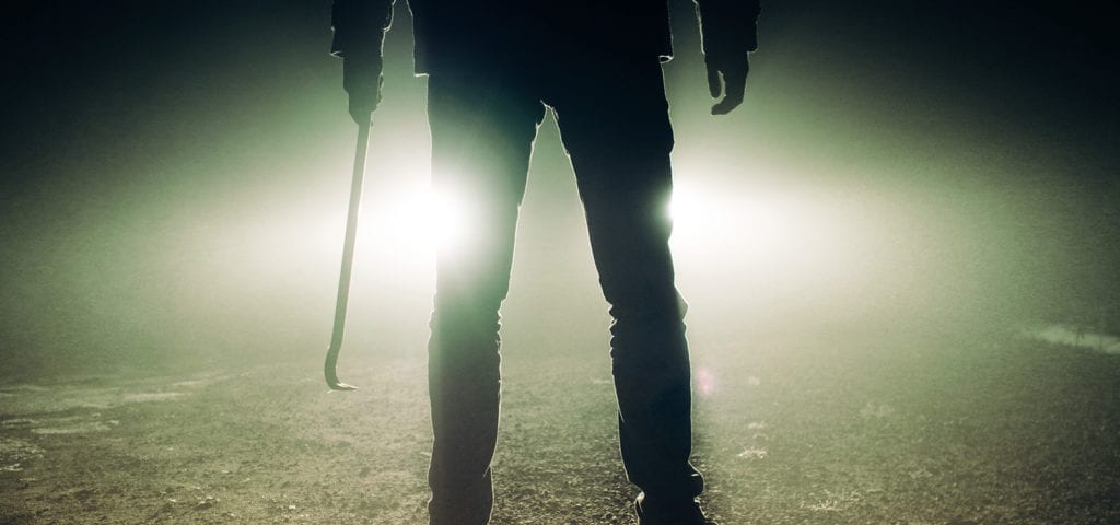 A man stands in front of car headlights while holding a crowbar.