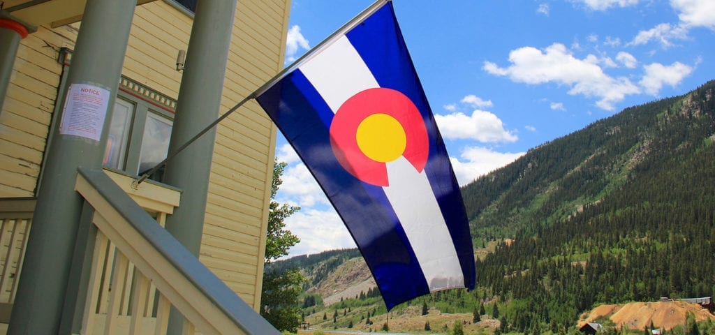 The Colorado state flag hangs off of the front porch of a large house located in the Rocky Mountains.